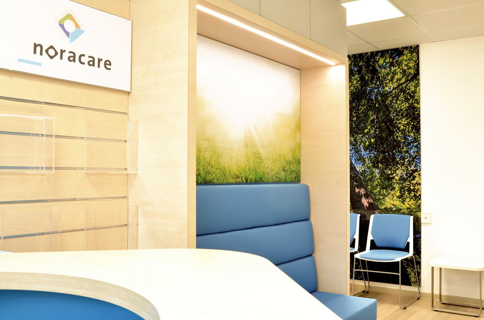 Empfangsbereich von noracare mit hellblauer Sitzbank, modernem Design und Hintergrundbild von B&auml;umen und Sonnenlicht.