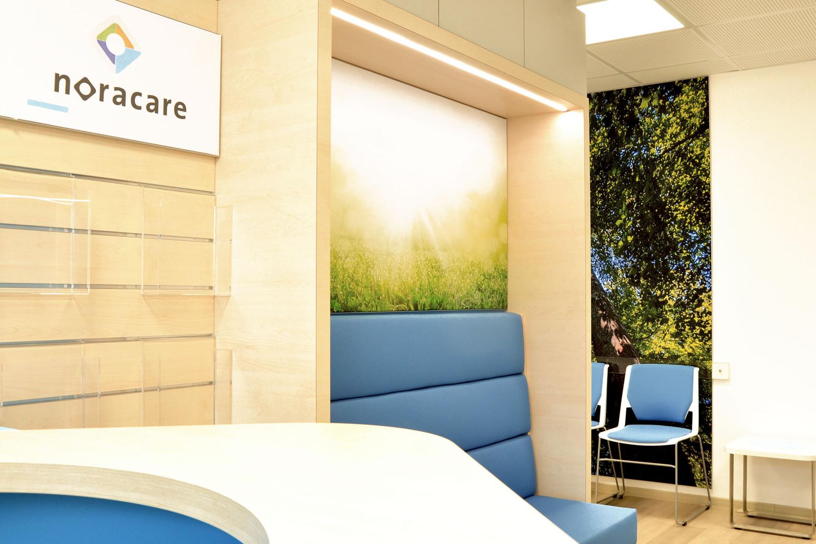 Empfangsbereich von noracare mit hellblauer Sitzbank, modernem Design und Hintergrundbild von B&auml;umen und Sonnenlicht.