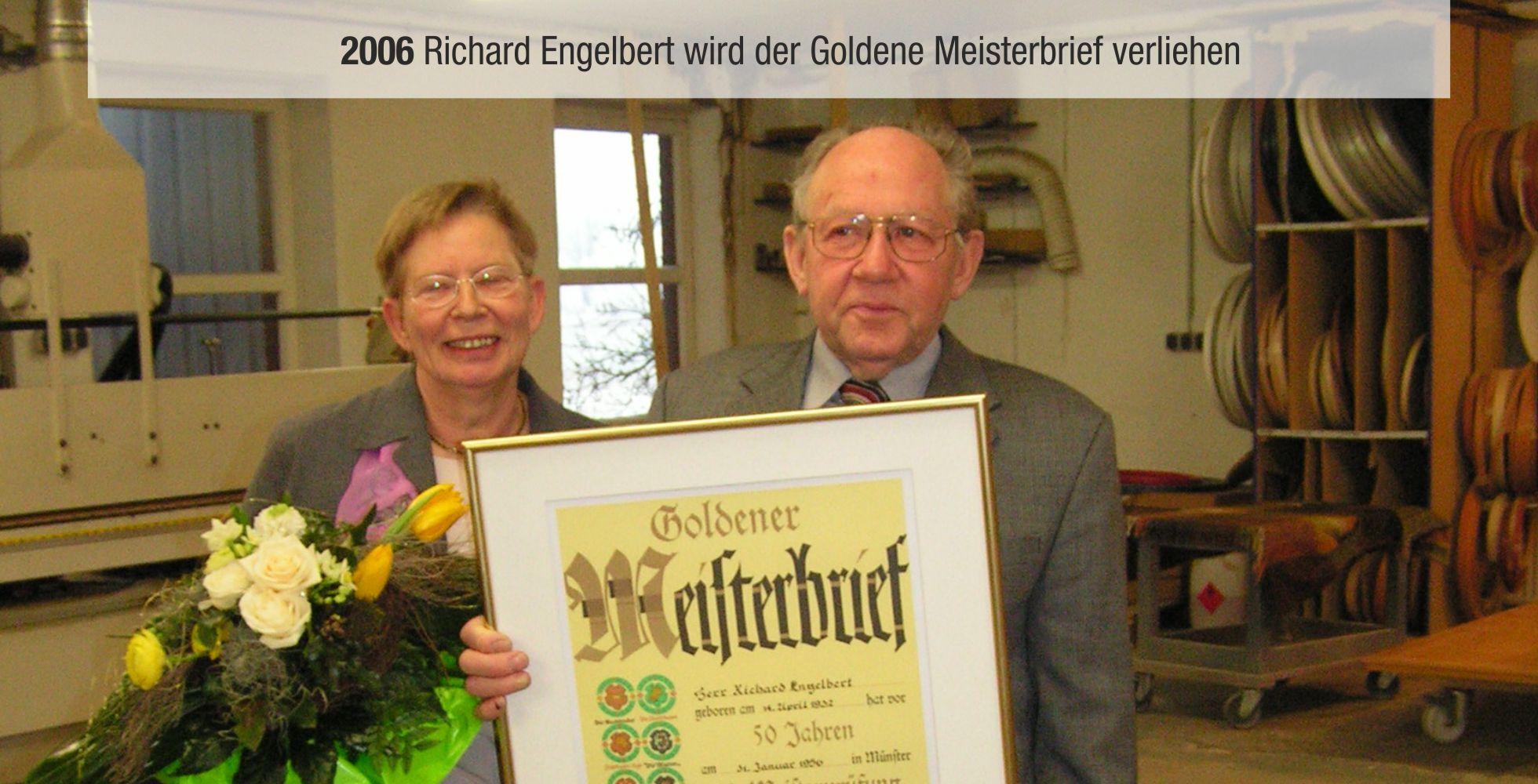 Richard Engelbert h&auml;lt den Goldenen Meisterbrief, w&auml;hrend eine Frau mit Blumen neben ihm steht. Hintergrund: Werkstattumgebung.