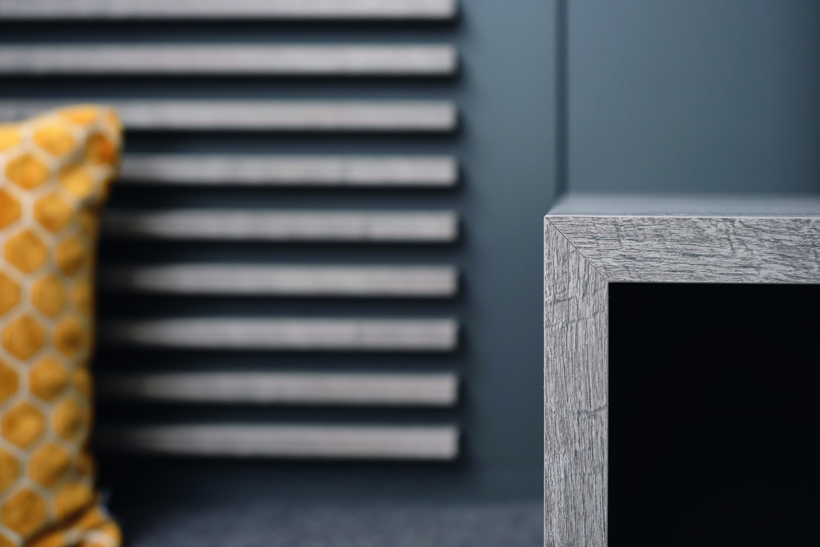 Nahaufnahme einer grauen Möbelkante mit verschwommener gelber Dekokissen und gestreifter Wand im Hintergrund.